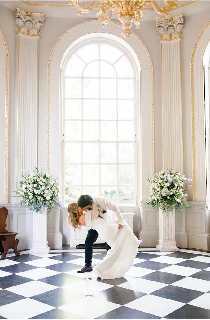 A newly married couple kissing between pastel flower arrangements beneath a grand arched window