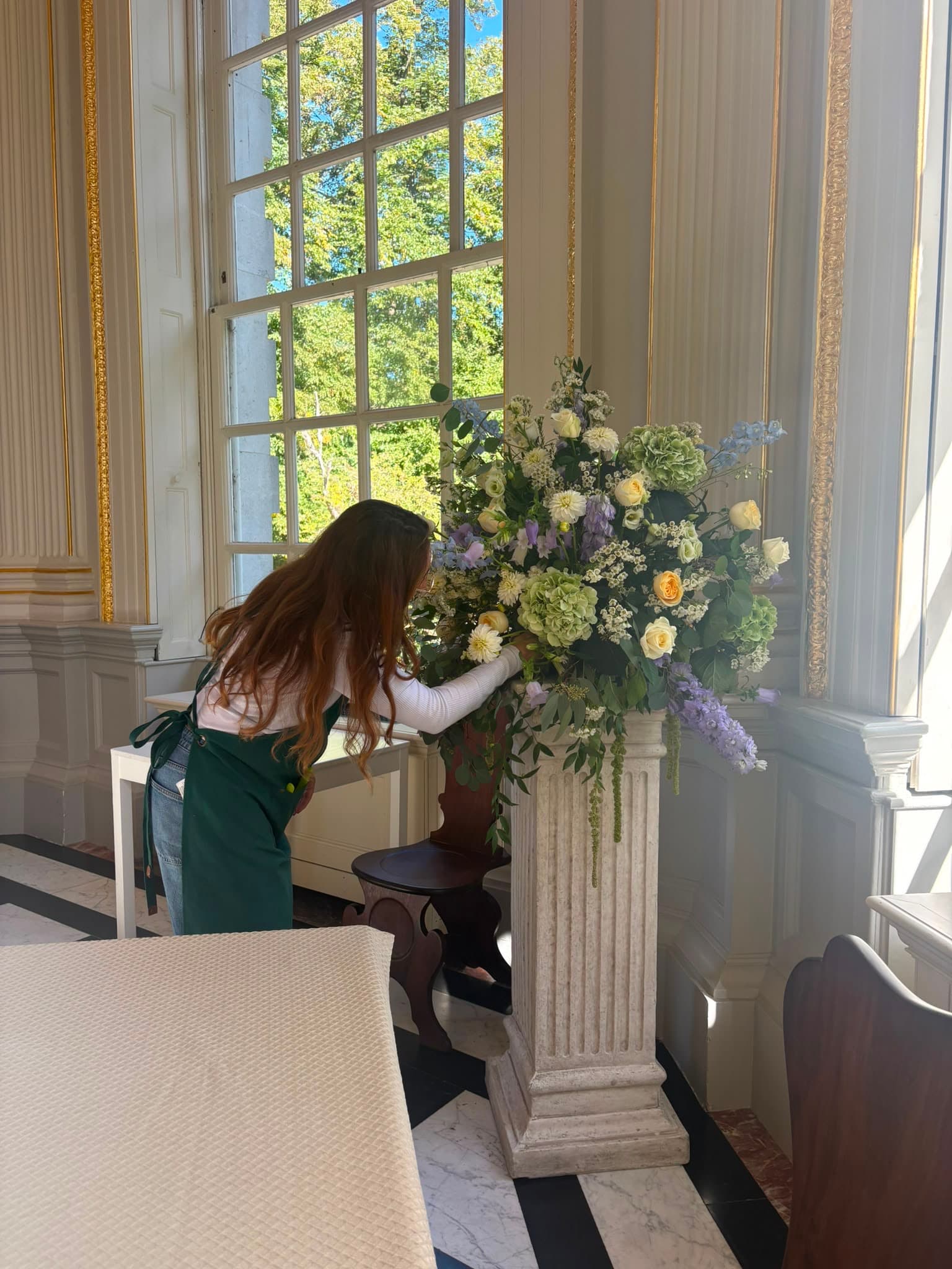 A florist arranging pastel wedding flowers on a stone pedestal in a sunlit room
